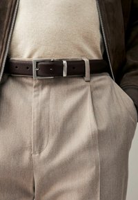 Brown leather belt with a silver rectangular buckle, worn over light beige tailored trousers with pleats and a cream-colored top underneath.