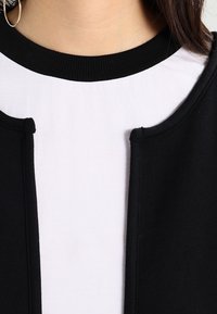 Svart och vit topp med ribbad halsringning. Har en rundad krage och ett kontrasterande svart ytterlager som ramar in en vit mittpanel.