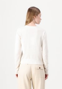 Maglione color crema con maniche lunghe, scollo rotondo, polsini e orlo a costine. Tessuto morbido; indossato sopra pantaloni beige. Vista posteriore.
