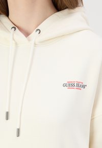 Krēmkrāsas kapuce ar auklu, kurai ir sarkans un melns logo. Mīksta tekstūra, minimālisma dizains, tuvplāns uz kakla zonu.