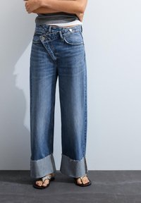 Personne portant un jean bleu à jambes larges avec des revers hauts roulés, un haut gris et des sandales noires avec des brides en cuir de serpent, debout contre un mur clair.