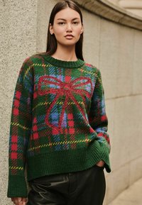 Zelený pletený sveter s károvaným vzorom v červenej, modrej a žltej farbe. Má veľký červený trblietavý motív mašle na prednej strane a rebrované manžety.