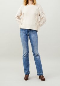 Maglione crema lavorato a maglia con motivi testurizzati e frange, abbinato a jeans a zampa d'elefante blu chiaro e scarpe con platform marroni.