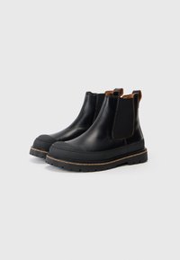 Bottes Chelsea en cuir noir avec semelles en caoutchouc épaisses, panneaux latéraux élastiques et tirettes marron, posées sur un fond blanc.