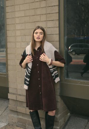 Bordeaux corduroy shirtjurk met korte mouwen, knopen aan de voorkant, en een zwart en crème-gepatteerd gebreid vest over de schouders gedrapeerd. Zwarte kniehoge laarzen.