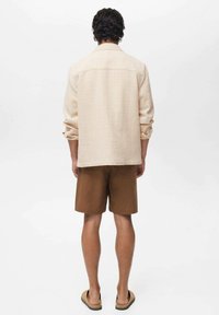 Light beige relaxed-fit shirt with rolled sleeves and a back yoke, paired with brown shorts. Model is wearing dark sandals.