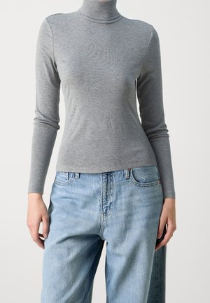 Person iført en tætsiddende grå rullekrave-trøje kombineret med lyseblå, højttaljede jeans mod en ensfarvet baggrund.