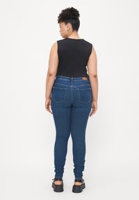 Dark blue skinny jeans with a high waist, featuring back pockets and a tag. Paired with a black sleeveless top and chunky black sandals.