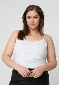 Witte camisole-top met dunne bandjes, met een ronde halslijn en zachte stof. Gekocht met zwarte broek, handen zichtbaar op de taille.