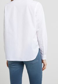 Camicia bianca a maniche lunghe, in tessuto leggero, taglio dritto con spacchi laterali; abbinata a jeans blu con dettagli sulle tasche.