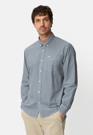 Homme aux cheveux bouclés portant une chemise à carreaux bleue et blanche boutonnée et un pantalon beige, debout les mains dans les poches.