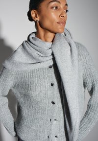 Cardigan tricoté gris avec nervures verticales, boutons noirs, et une écharpe grise assortie drapée lâchement autour du cou. Matière douce et texturée.