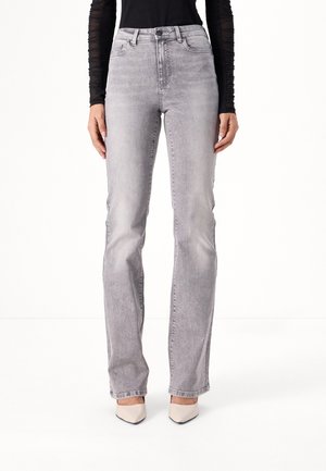 Kvinde iført lysegrå jeans med lige ben, sort tætsiddende top med lange ærmer og hvide spidse hæle, stående mod en ensfarvet baggrund.