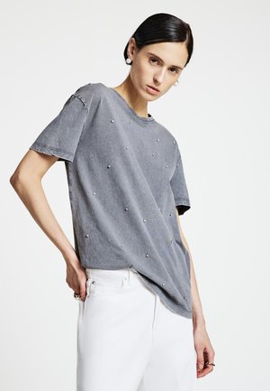 Graues Baumwoll-T-Shirt mit lockerem Schnitt, Rundhalsausschnitt und dekorativen silbernen Nieten auf der Vorderseite. Kombiniert mit weißen Hosen.