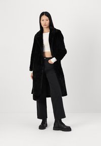 Abrigo de piel sintética negro con un collar ancho, mangas largas y un frente abierto, combinado con un top corto blanco y pantalones de pierna ancha negros.