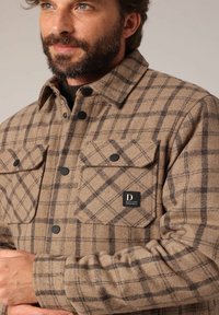 Bruine geruite flanellen jas met zwarte knopen en twee borstzakken. Heeft een textuurstof met een zichtbaar logo label en kraag.