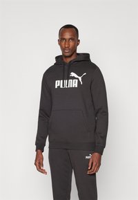 Schwarzer Hoodie aus weichem Stoff mit Kängurutasche, Kapuze mit Kordelzug und weißem Branding. Kombiniert mit passenden Sweatpants.