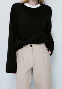 Persona bionda che indossa un maglione nero largo sopra una t-shirt bianca infilata in pantaloni beige a vita alta, con le mani in tasca.