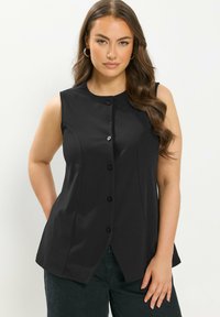 Mujer con cabello largo y ondulado, vistiendo una blusa sin mangas negra abotonada y pantalones negros, de pie con una mano en el bolsillo contra un fondo blanco.