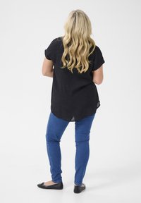 Černý krátký rukáv s volným střihem, modré skinny džíny a černé baleríny. Dlouhé vlnité vlasy viditelné zezadu. Jednoduché bílé pozadí.