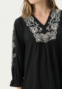 Schwarzes Kleid mit V-Ausschnitt-Design, verziert mit weißen gestickten Blumenmustern am Ausschnitt und an den Ärmeln. Es hat eine geraffte Taille und Glockenärmel.