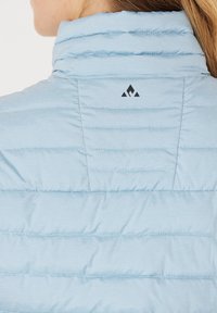 Lyseblå, pufmærket jakke med høj krave, vandret syning og et lille sort logo på ryggen. Blød, glat tekstur.