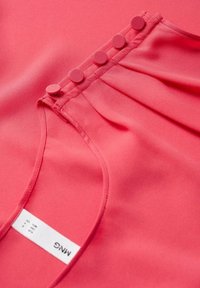 Blouse rose en tissu lisse, ornée de cinq boutons recouverts de tissu le long de l'encolure et d'une étiquette avec le logo de la marque et les instructions d'entretien.