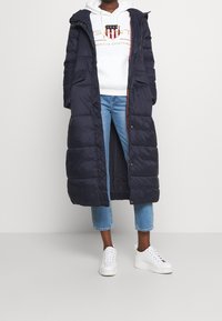 Giubbotto imbottito blu navy con cappuccio, dal design lungo, texture trapuntata e due tasche, indossato sopra un hoodie bianco e jeans chiari.