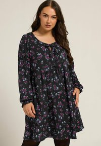 Musta kukalllinen mekko, jossa on violetteja ja sinisiä kukkia, pitkät hihat, pyöreä pääntie ja etuosan solmimisdetail; pehmeä, tekstuuri kangas.