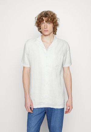 Camicia bianca di lino a maniche corte con colletto a scollo, chiusura con bottoni e taschino sul petto, abbinata a pantaloni in denim blu.