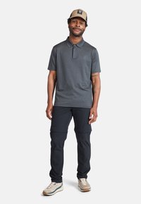 Polo shirt grigio con colletto, abbinato a pantaloni cargo neri e sneakers beige. Il cappello è beige con una patch del logo. Outfit casual sportivo.