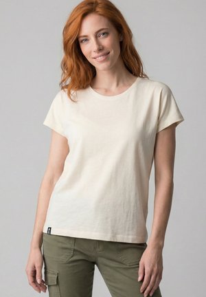 Red-haired woman wearing a plain cream short-sleeve t-shirt and olive green cargo pants, standing and smiling against a gray background.