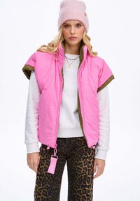 Prošívaná vesta bez rukávů v jasně růžové barvě s olivově zelenými lemy; doplněná bílým svetrem a kalhotami s leopardím vzorem; obsahuje visačku.