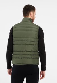 Grüne gepolsterte Weste mit hohem Kragen, mit horizontalen Nähten, leichtem Material und einem eleganten Rückendesign. Über einem schwarzen Langarmshirt getragen.