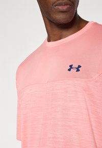 Αθλητική μπλούζα σε ανοιχτό ροζ χρώμα με υφή σχεδίου, στρογγυλή λαιμόκοψη και λογότυπο Under Armour σε σκούρο μπλε στο στήθος.