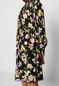 Abito floreale con sfondo nero, caratterizzato da fiori colorati in rosa, giallo e bianco. Maniche lunghe e silhouette morbida con gonna arricciata.