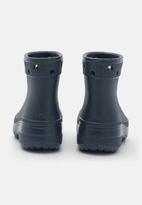 Une paire de bottes en caoutchouc noires jusqu'à la cheville avec des semelles texturées et des poignets à motifs, vues de l'arrière sur un fond blanc.