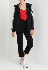 Svart zip-up hoodie med vita ränder på ärmarna, röd linne, svarta cropped byxor och vita sneakers. Rena linjer, avslappnad design.