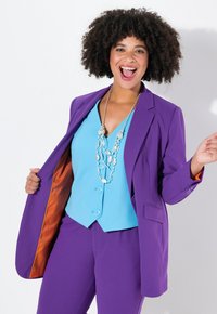 Blazer viola e pantaloni abbinati indossati con una canotta blu senza maniche. Presenta una fodera interna arancione e una collana d'argento.