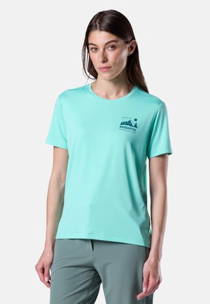Donna che indossa una maglietta Rossignol turchese a maniche corte con logo montagna e pantaloni verde salvia, in piedi contro uno sfondo chiaro e uniforme.