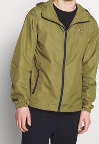 Olivgrön lättviktsjacka med huva, elastiska ärmslut, marinblå dragkedja och sidofickor; slät yta och avslappnad design.