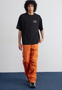 Juodos oversized marškinėliai su XTC logotipu, derinami su ryškiais oranžiniais cargo kelnėmis, turinčiomis kišenes ir laisvą siluetą. Juodi ir baltai sportiniai bateliai.