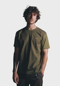 Olijf groene t-shirt met korte mouwen, een ronde halslijn en textuuraccenten op de schouders. Heeft een klein olifantenlogo op de borst.