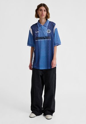 Maglia sportiva azzurro chiaro con motivo a rombi blu scuro, dettagli bianchi e testo del logo nero. Abbinata a jeans neri larghi.