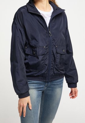 Femme portant une veste bleu marine zippée avec des poches avant à rabat et des poignets élastiques, associée à un jean bleu clair et un t-shirt blanc.