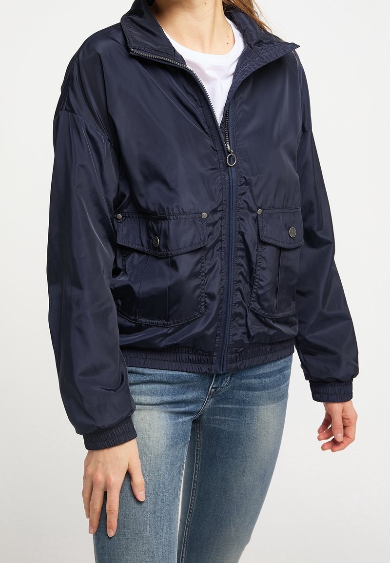 Femme portant une veste bleu marine zippée avec des poches avant à rabat et des poignets élastiques, associée à un jean bleu clair et un t-shirt blanc.