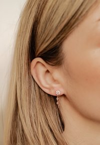 Ohrringe aus Roségold mit einem blumigen Ohrstecker, kleinen Ketten und zarten Blattausbesserungen, vor einem hellen Hintergrund.