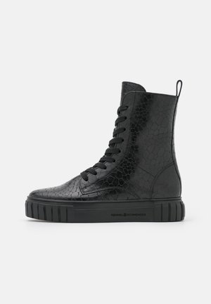 Botte montante noire avec motif crocodile texturé, semelle épaisse en caoutchouc, lacets à l'avant et languette à l'arrière, vue latérale sur fond blanc.