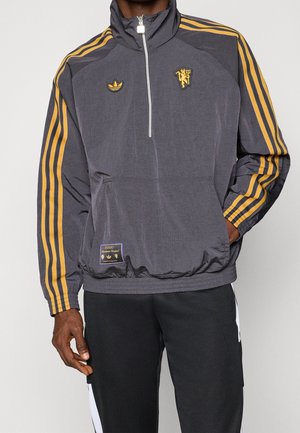 Hombre con chaqueta gris oscuro con media cremallera y franjas naranjas en las mangas, con los logotipos de Manchester United y Adidas, y un bolsillo delantero tipo canguro.