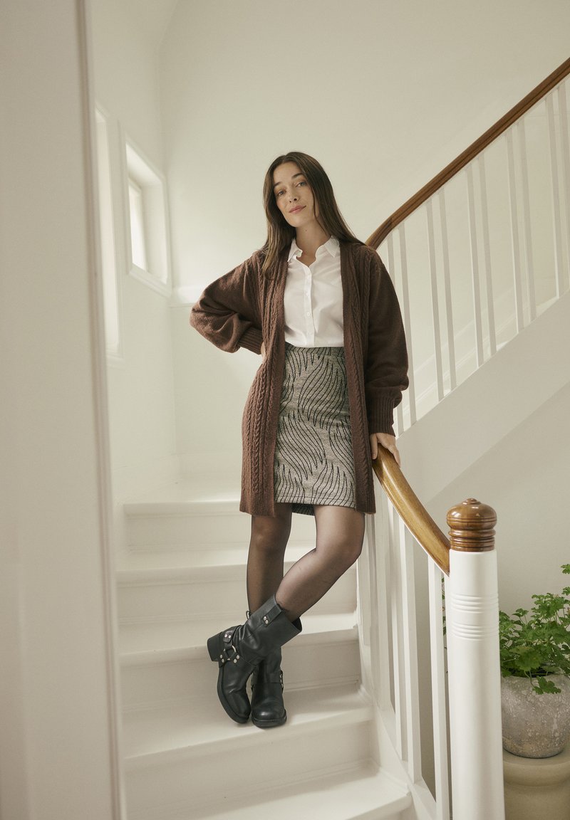 Brown knitted cardigan over a white shirt, patterned skirt with wave design, black ankle boots, standing on a staircase with wooden railing.